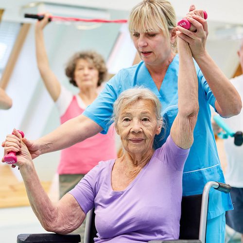 Rehaübungsleiterin unterstützt eine ältere Reha-Patientin beim Ausführen der Übung für den medizinischen Rückensport